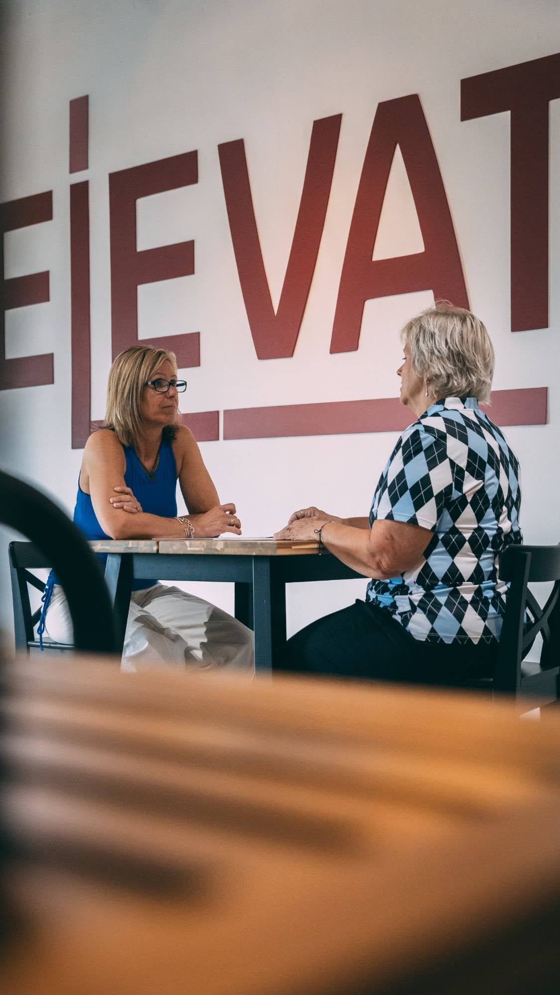 Jaime Hughes and a client in a relaxed conversation at a table beneath the large ELEVATE wordmark in the studio