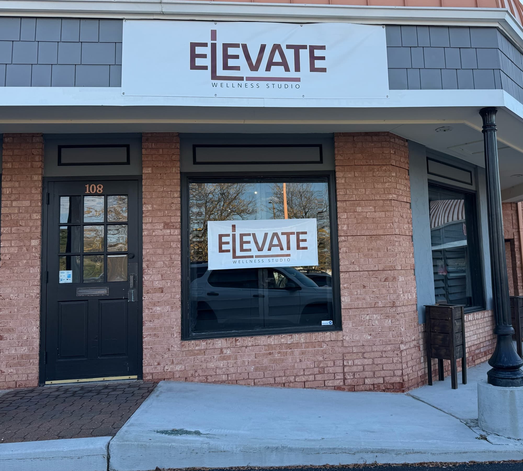 The Ballwin entrance of Elevate Wellness Studio: a black door numbered 108 beside a brick storefront, with an ELEVATE Wellness Studio sign above and another in the window