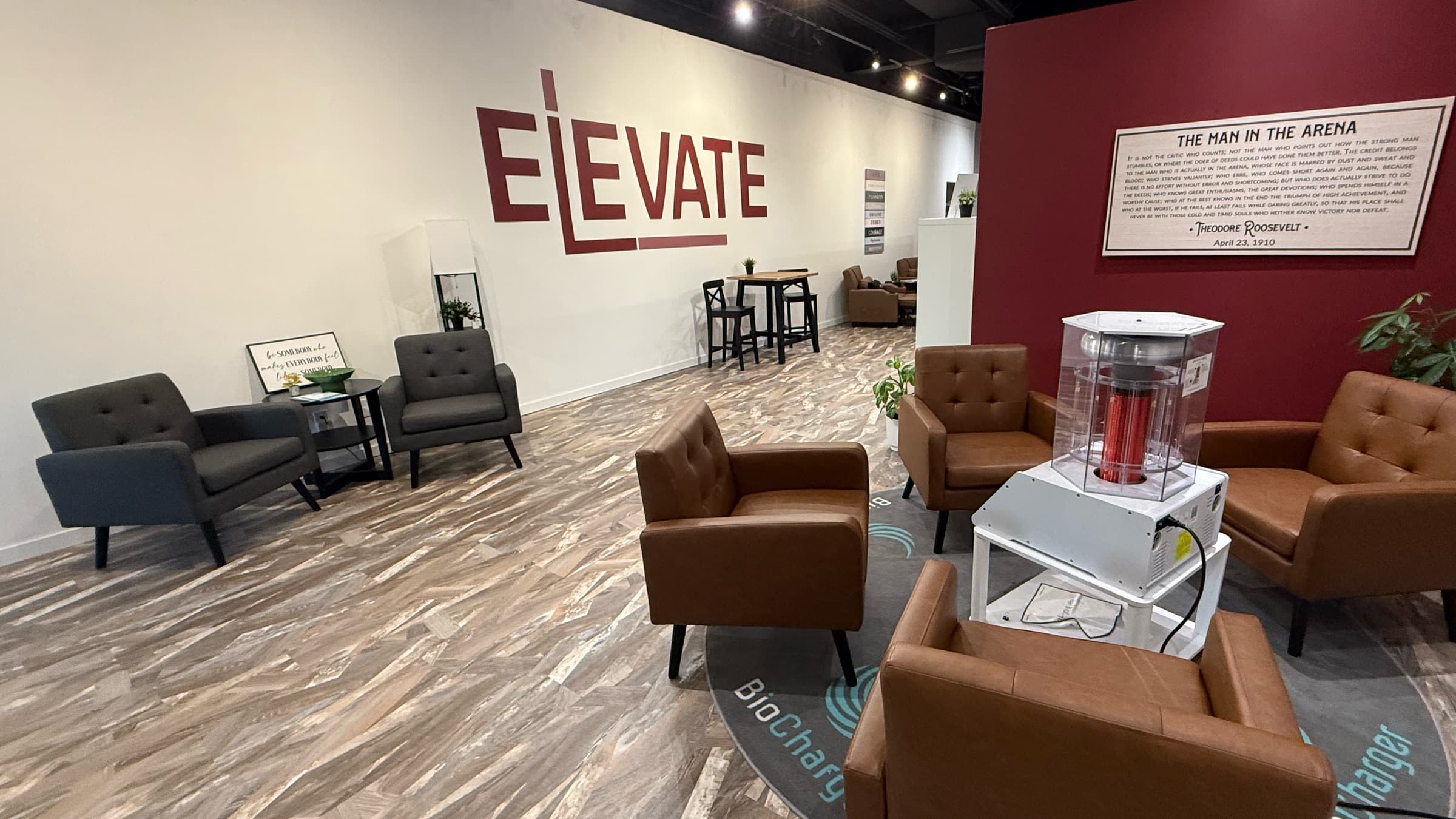 Inside Elevate Wellness Studio: the maroon ELEVATE logo on a long white wall, lounge seating, and leather armchairs gathered around the BioCharger