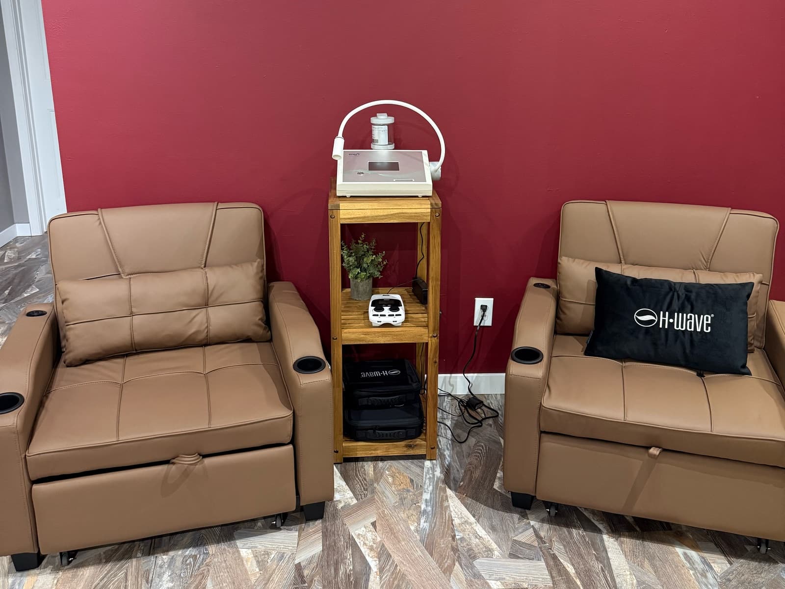 Two leather recliners on either side of a wooden cart holding the NanoVi and H-Wave devices, against a crimson wall at Elevate Wellness Studio