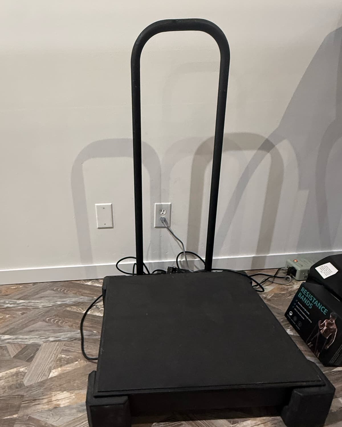 The whole-body vibration plate at Elevate Wellness Studio: a low black platform with a tall U-shaped handle, against a white wall